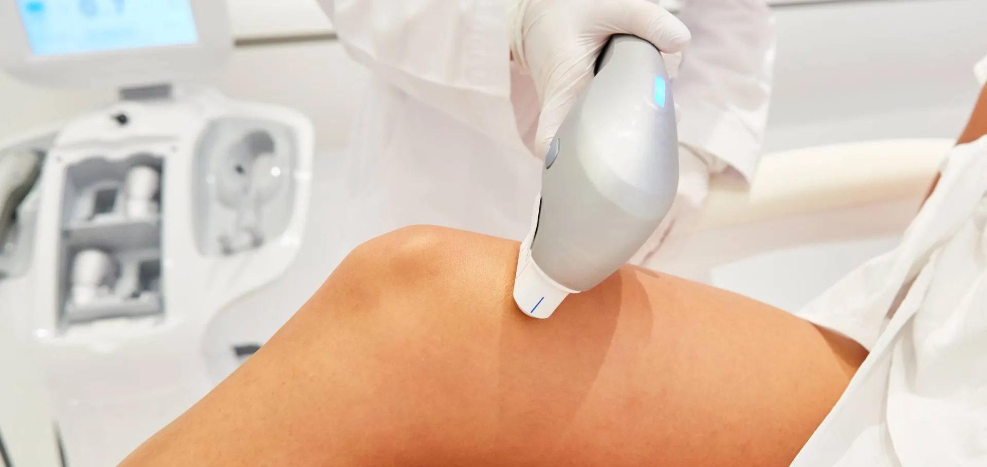 Aplicação de ultrassom macrofocado na perna da paciente, profissional com luva conduzindo o aplicador junto ao equipamento, ambiente clínico claro com tela azulada, fotografia de tratamento corporal profissional